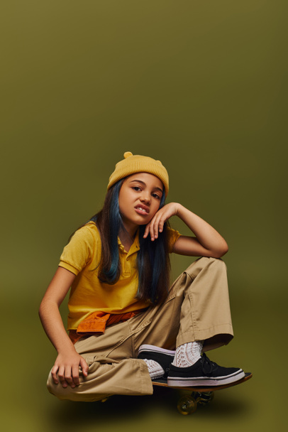 Disgusted and fashionable preteen girl with dyed hair wearing yellow hat and urban outfit while looking at camera and sitting on skateboard on khaki background, girl in urban streetwear concept - Photo, Image