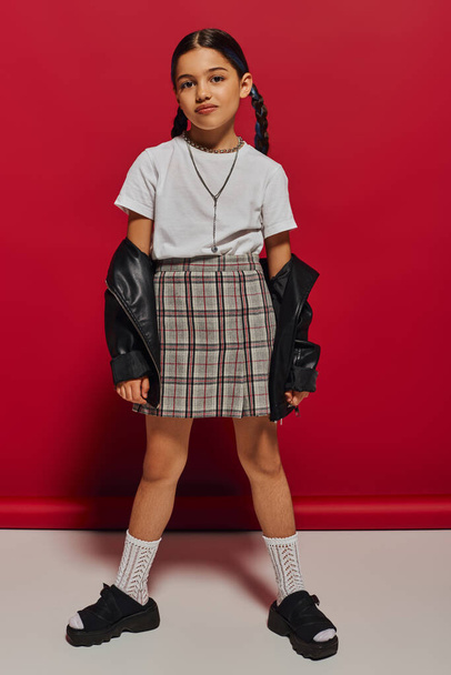 Full length of trendy brunette preteen girl with hairstyle posing in plaid skirt and leather jacket while looking at camera and standing on red background, stylish preteen outfit concept - 写真・画像