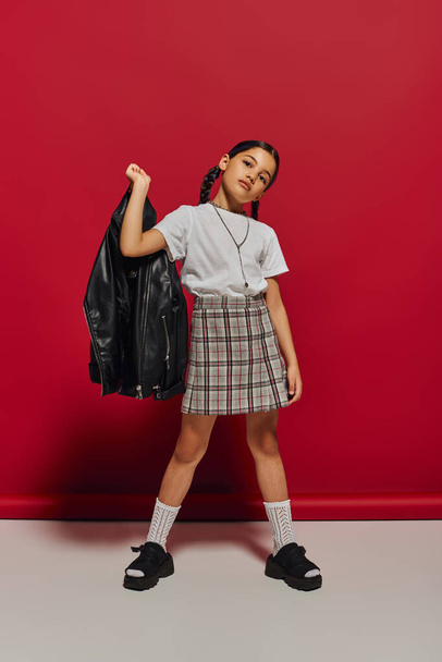 Brunette preadolescent girl with hairstyle holding leather jacket while posing in checkered skirt and looking at camera while posing on red background, stylish preteen outfit concept - Foto, afbeelding