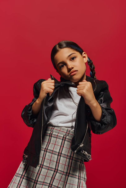 Portrait of fashionable preadolescent girl with hairstyle posing in checkered skirt while holding leather jacket and looking at camera isolated on red, stylish preteen outfit concept - Foto, afbeelding