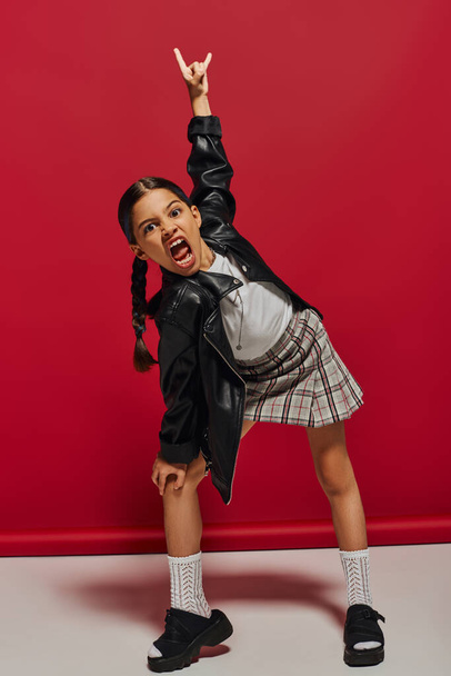 Mad and trendy preteen girl with hairstyle wearing leather jacket and plaid skirt looking at camera and showing rock gesture while standing on red background, stylish preteen outfit concept - Foto, Imagem