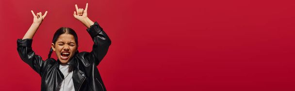 Opgewonden en modieus preadolescent meisje met moderne kapsel dragen lederen jas en tonen rock gebaar geïsoleerd op rood, meisje met koele en eigentijdse look, banner met kopieerruimte - Foto, afbeelding
