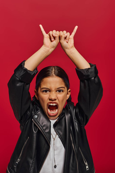 Portret van gekke en modieuze pre-adolescente meisje in leren jas schreeuwen op de camera en tonen rock gebaar terwijl staande geïsoleerd op rood, meisje met koele en eigentijdse look - Foto, afbeelding