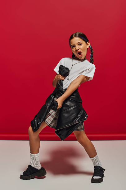 Excited and trendy preteen girl with hairstyle wearing t-shirt and plaid skirt while holding leather jacket and standing on red background, hairstyle and trendy accessories concept - 写真・画像