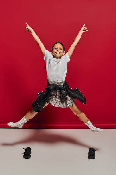 Cheerful and trendy preteen girl in plaid skirt, t-shirt and leather jacket pointing with fingers while jumping and looking at camera on red background, hairstyle and trendy accessories concept - Fotó, kép