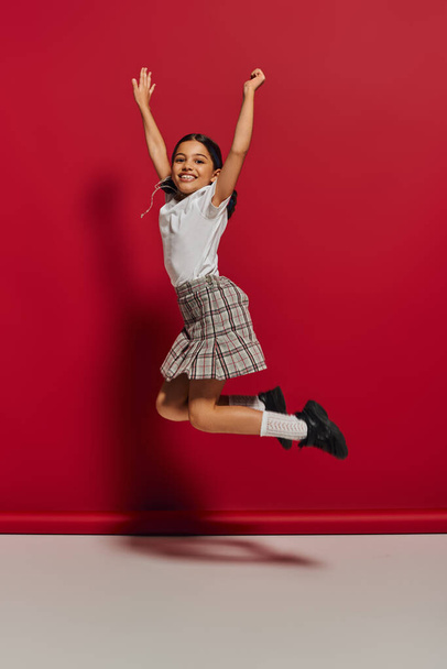Positive and trendy brunette preteen girl in t-shirt and checkered skirt jumping while having fun and looking at camera on red background, hairstyle and trendy accessories concept - Foto, Imagem