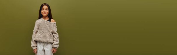 Trendiges vorpubertierendes brünettes Mädchen in Strickpullover und Herbstoutfit, das im Stehen und isoliert posiert auf grünem, entspanntem Herbstambiente in die Kamera blickt  - Foto, Bild