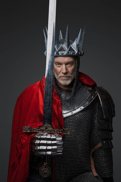 Porträt eines charmanten, reifen Königs mit grauem Bart, der eine Stahlkrone und schwere Rüstung trägt, wobei eine Schulter von einem roten Mantel bedeckt ist und ein Schwert vor einem grauen Hintergrund hält. - Foto, Bild