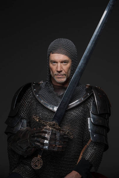 Pewny siebie portret dojrzałego króla z szarą brodą, noszącego ciężką zbroję, trzymającego miecz na szarym tle - Zdjęcie, obraz