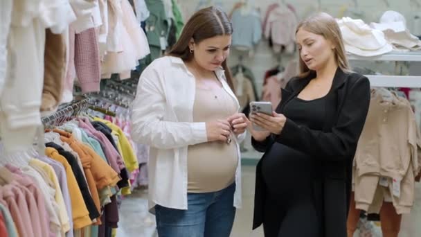 Dos mujeres embarazadas felices comprando ropa en la tienda del bebé, mire el producto en el teléfono, eligiendo una cintura - Imágenes, Vídeo