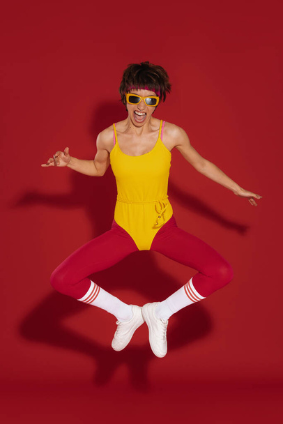 Happy young woman in retro styled sportswear jumping against red background - Foto, immagini