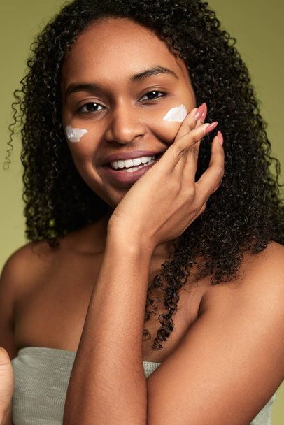 Cheerful Hispanic female with curly hair touching cheek while applying moisturizing cream on face during skincare routine looking at camera against green background - Zdjęcie, obraz