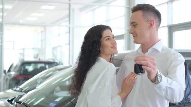 Happy married couple owners of a new car. They are in a car dealership and show the keys. Car business concept - Кадры, видео