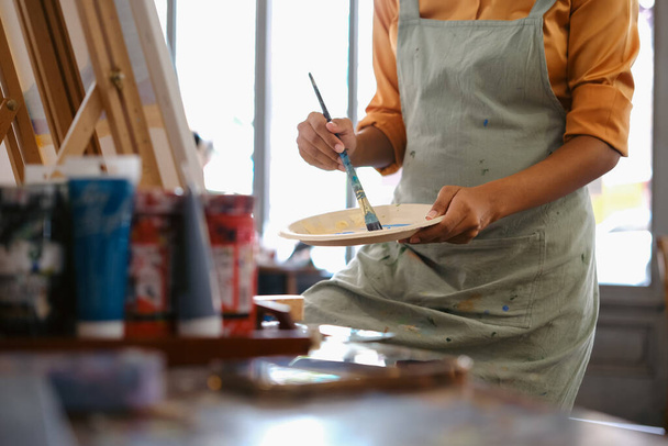 Καλλιεργημένη εικόνα γυναικείας καλλιτέχνιδας που στέκεται μπροστά από ένα καβαλέτο και βουτάει πινέλο στην παλέτα χρωμάτων. - Φωτογραφία, εικόνα