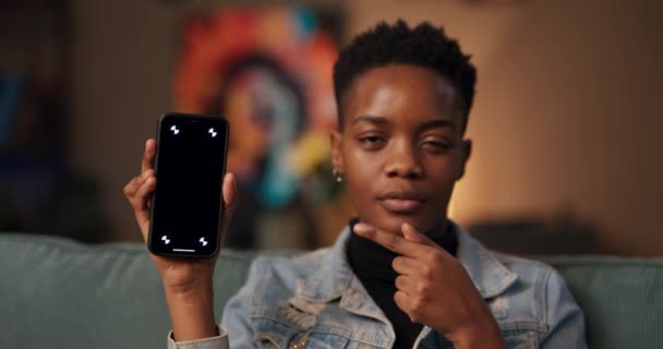 熱狂的な技術レビューの影響力アフリカの女性は、カメラにそれを紹介し、彼女の洞察力と推奨事項を彼女の聴衆と共有し、手に暗い画面で彼女の携帯電話を保持しています. - 映像、動画