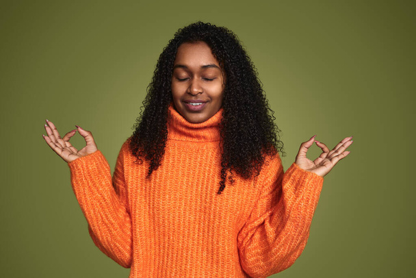 Calm black female in orange sweater and with curly hair holding Gyan mudra and meditating to relief stress on green background - Fotó, kép