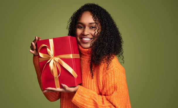 Cheerful african american girl in trendy orange sweater with curly hair, smiling and holding a big gift box over green background - Photo, image