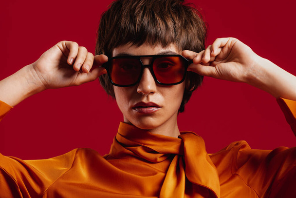 Portrait of fashionable young short hair woman adjusting her trendy glasses against red background - Fotó, kép