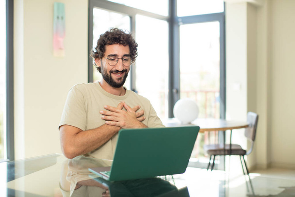 young adult bearded man with a laptop feeling romantic, happy and in love, smiling cheerfully and holding hands close to heart - Foto, Imagen