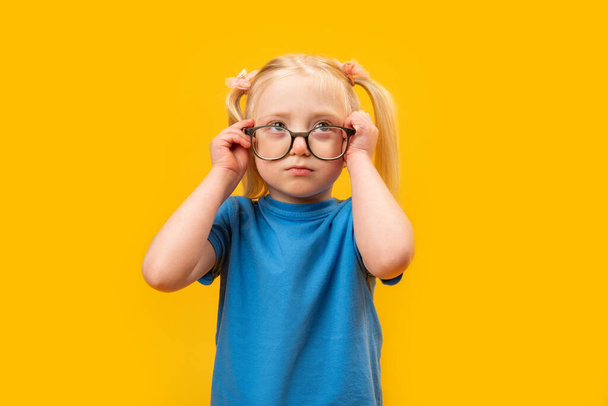 Elementary schoolgirl with two ponytails wears blue T-shirt and glasses looks up. Studio portrait of child, yellow background. - Фото, изображение