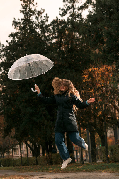 Fröhlicher Student in schwarzem Mantel mit durchsichtigem Regenschirm springt über die Gasse des Herbstparks. Üppiges Haar des Mädchens verdeckt Gesicht. - Foto, Bild