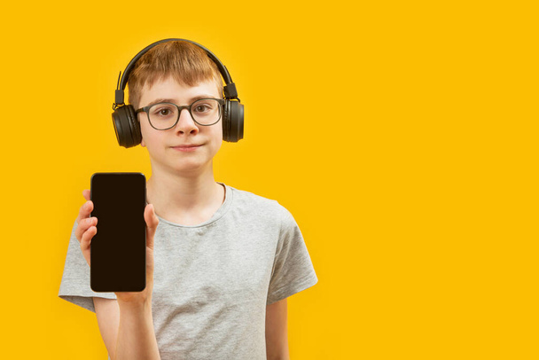 Teenage brunet boy with glasses wears headphones and holds smartphone with blank black screen. Copy space. Mock up - Fotó, kép