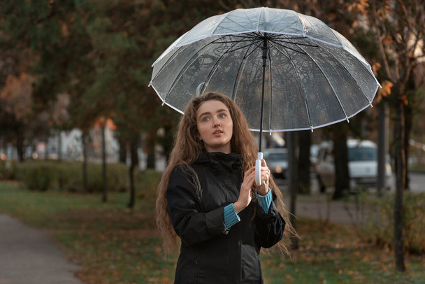 Gyönyörű lány barna hajjal sétál az őszi parkban. Fiatal nő átlátszó esernyő látni esik. - Fotó, kép