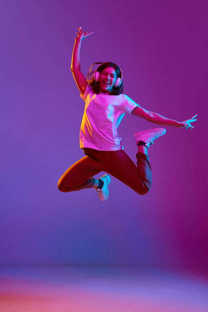 Full-length portrait of young, happy, smiling girl in casual clothes as headphones jumping over gradient purple background in neon light. Concept of emotions, youth, feelings, fashion, lifestyle, ad - Zdjęcie, obraz