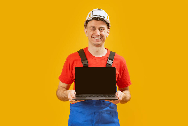 Ein Mann mittleren Alters trägt einen Schutzhelm, in dessen Hand ein Laptop mit leerem Bildschirm auf gelbem Studiohintergrund liegt. Kopierraum, Attrappe - Foto, Bild