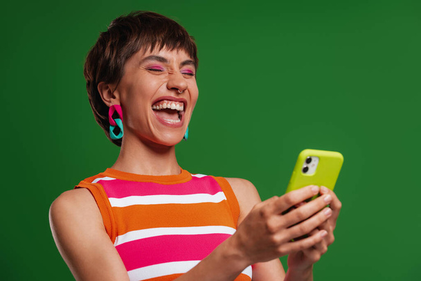 Ενθουσιασμένη νεαρή γυναίκα κρατώντας έξυπνο τηλέφωνο και χαμογελώντας ενώ στέκεται πάνω από το πράσινο φόντο - Φωτογραφία, εικόνα