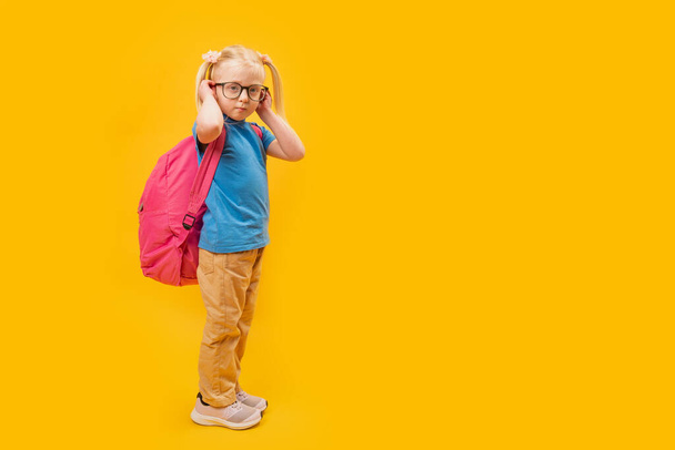 Ritratto a figura intera di Serious bambina bionda che indossa lo zaino per la scuola, sfondo giallo. Copia spazio, mockup. - Foto, immagini