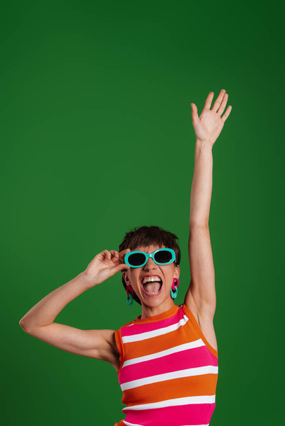 Happy young woman in trendy glasses gesturing while standing against green background - Foto, imagen