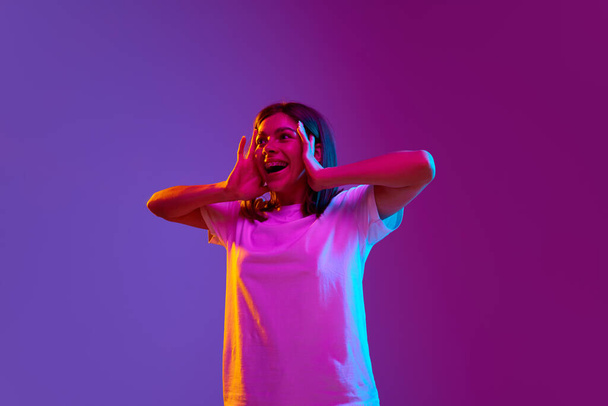 Portrait of excited, happy, smiling young girl standing with positive shocked face on gradient purple background in neon light. Surprise. Concept of emotions, youth, feelings, fashion, lifestyle, ad - Zdjęcie, obraz