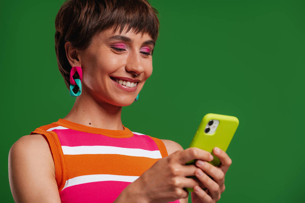 Attractive young woman using smart phone and smiling while standing against green background - Φωτογραφία, εικόνα