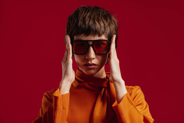 Portrait of stylish young short hair woman adjusting her trendy glasses against red background - Foto, imagen