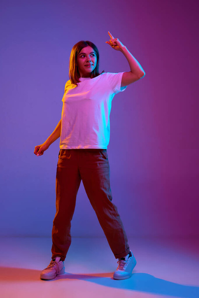 Full-length portrait of young girl in casual comfortable clothes standing with finger raised against gradient purple background in neon light. Concept of emotions, youth, feelings, fashion, lifestyle - Foto, Bild