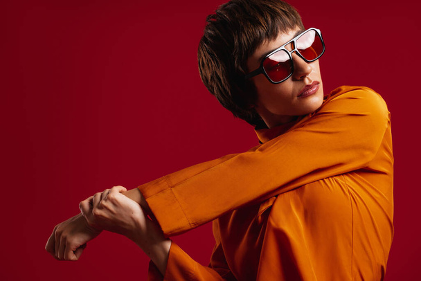 Fashionable young short hair woman in trendy glasses looking away against red background - Photo, Image