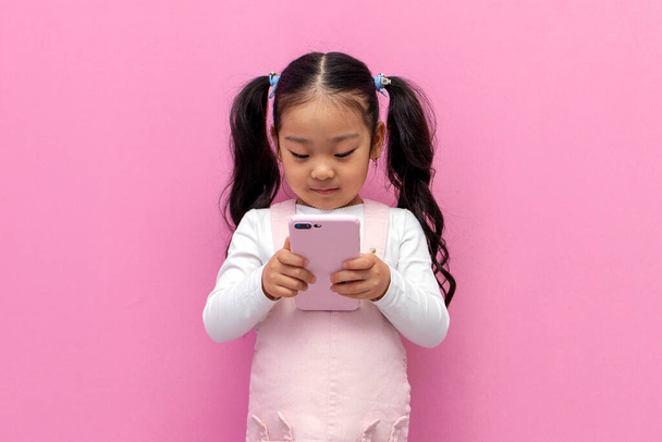little asian girl in pink sundress uses smartphone on pink isolated background, korean preschool child is typing on the phone - Foto, immagini