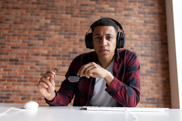 серйозний хлопець афроамериканський геймер в навушниках тримає окуляри в руках і дивиться на екран, чоловік сидить за комп'ютерним столом і знімає окуляри - Фото, зображення