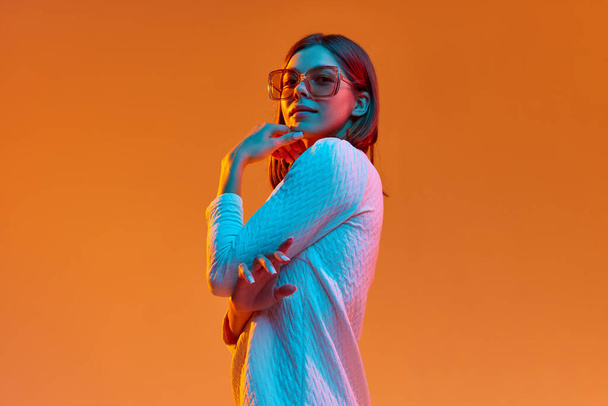 Portrait of young beautiful girl in casual clothes and trendy sunglasses posing against orange studio background in neon light. Concept of human emotions, youth, feelings, fashion, lifestyle, ad - Photo, image