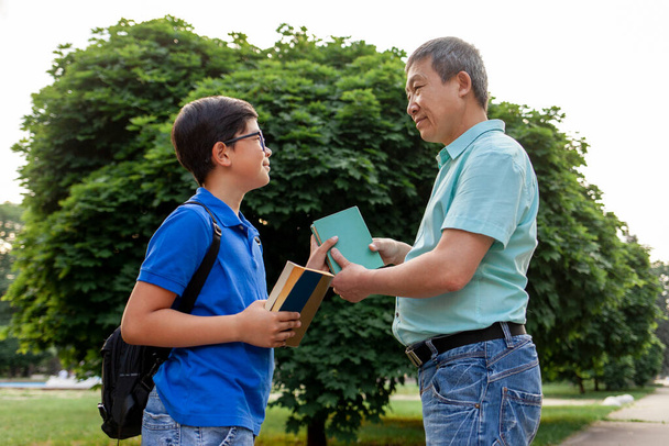 asiatischer alter Vater geht mit Sohn zur Schule und trägt Bücher, koreanischer Junge mit Rucksack und Brille geht im Sommer mit älterem Vater zur Schule, Großvater kümmert sich um Enkel - Foto, Bild