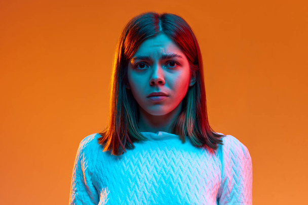 Close-up portrait of young girl standing with sad facial expression against orange studio background in neon light. Concept of human emotions, youth, feelings, fashion, lifestyle, ad - Zdjęcie, obraz