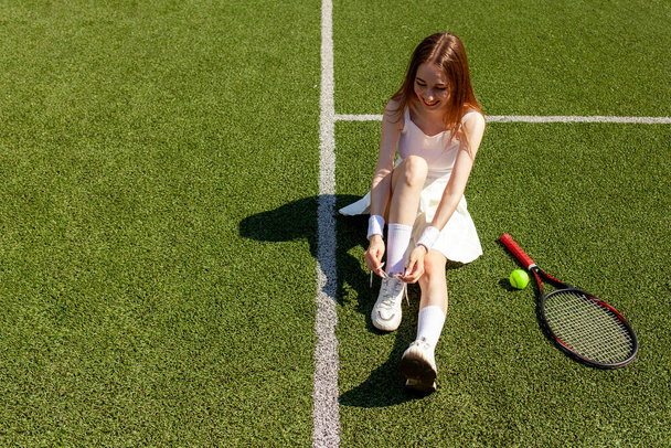 fiatal lány tenisz játékos fehér sport egyenruha köti a cipőfűzőjét tornacipő zöld pályán, női edző tenisz ütő, másolás helyet - Fotó, kép