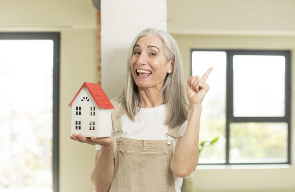Egy csinos idős nő boldog és izgatott zseninek érzi magát, miután rájött egy ötletre. egy ház modellel - Fotó, kép