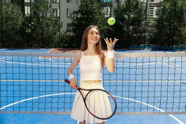 jong meisje tennisser in wit uniform met racket gooit de bal op blauwe veld buiten, vrouw tennis coach staat in de buurt van het tennisnet - Foto, afbeelding
