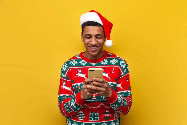 african american guy in christmas clothes and santa hat is using smartphone and smiling on yellow isolated background, man in sweater is typing online on phone for new year - Photo, Image