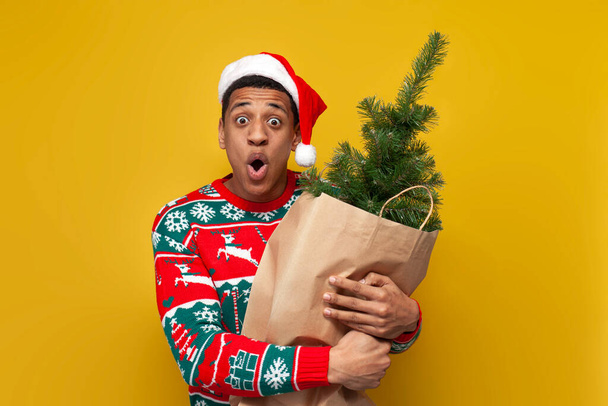 african american shocked guy in christmas clothes and santa hat is holding christmas tree in bag and is surprised on yellow isolated background, man in sweater is buying decorations for the new year - Photo, Image