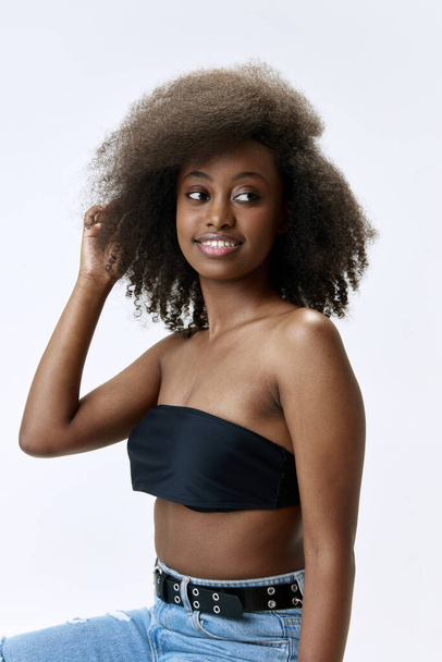 Beautiful, young, african woman with curly hair, smooth, healthy skin smiling, posing in jeans and top against white studio background. Concept of natural beauty, skin care, health, fashion, wellness - Photo, Image