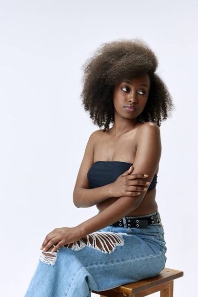 Portrait of beautiful, young, african woman with curly hair posing in top and jeans against white studio background. Concept of natural beauty, skin care, health, fashion, wellness, ad - Foto, Imagem