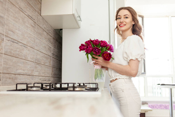 молодая девушка в белой одежде кладет букет красных тюльпанов в вазу дома на кухне, женщина кладет цветы в воду - Фото, изображение